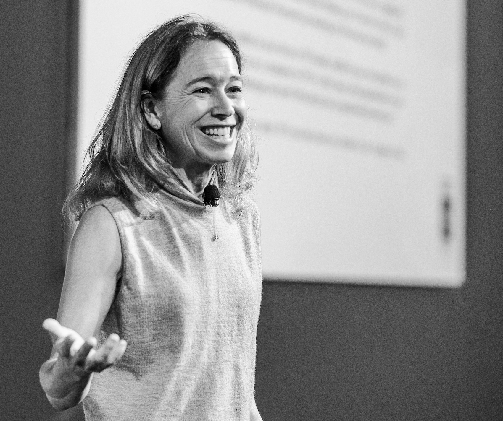 A black and white photograph of a smiling woman, Ali Levin, presenting in front of a large screen