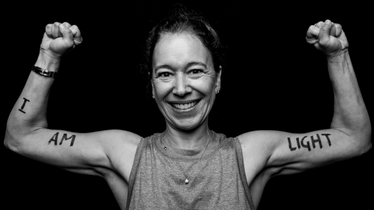 A black and white photo of a smiling woman raising both fists in the air. On her arms "I am light" is written in thick capital letters.